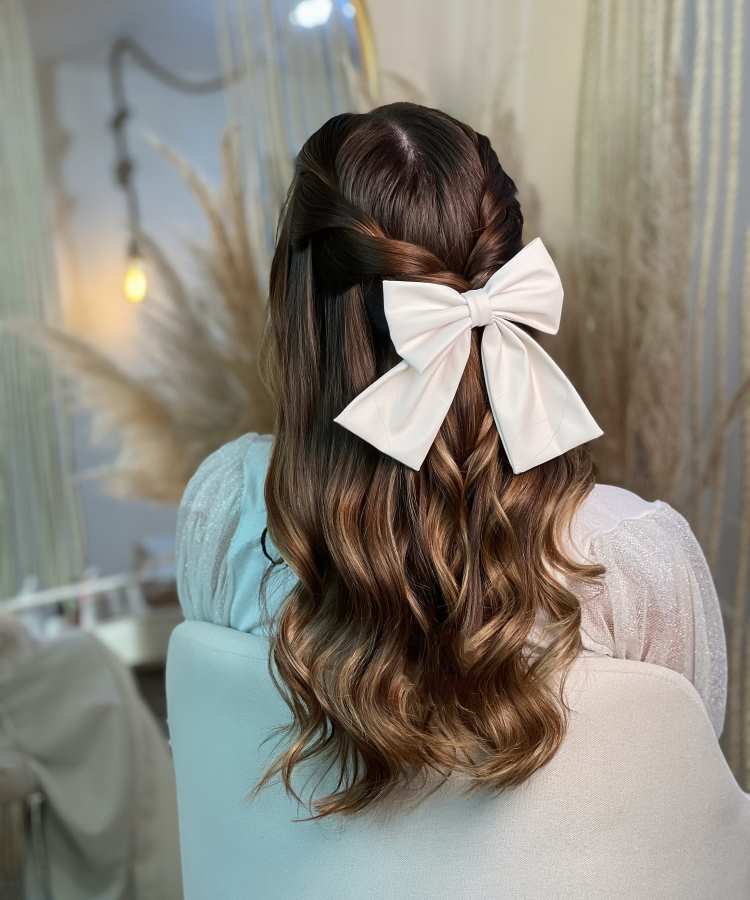 Cabelo castanho ondulado semipreso com tranças laterais, decorado com laço branco grande, em ambiente iluminado e delicado.