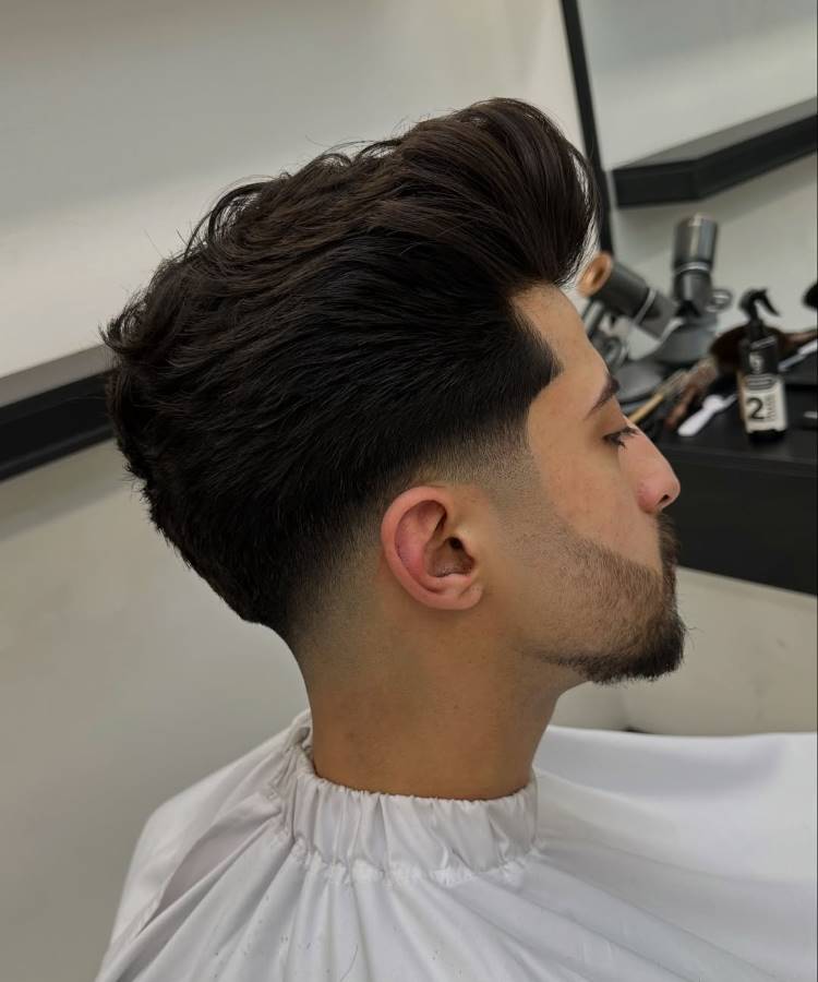 Um homem de perfil com um corte de cabelo pompadour e mid fade. O cabelo do topo é escuro, com bastante volume e penteado para trás. As laterais e a nuca têm um degradê.