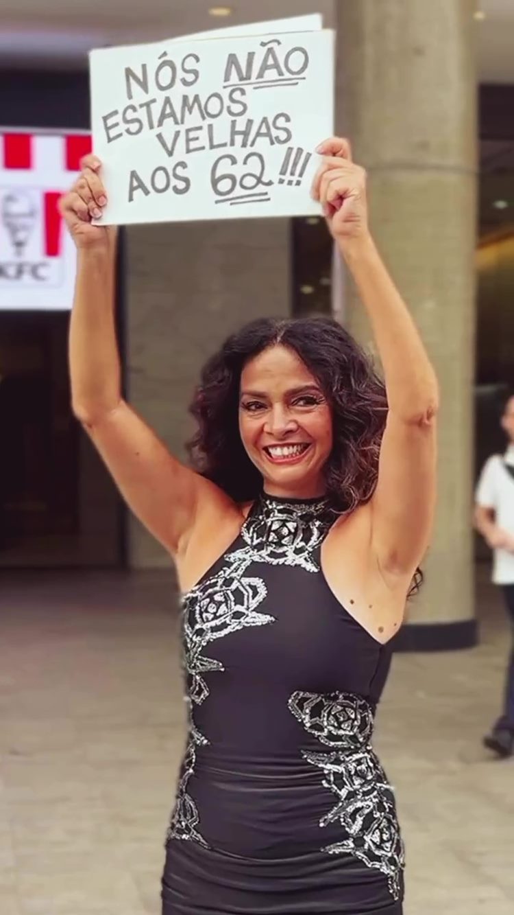 Claudia Ohana sorri enquanto ergue um cartaz com a frase “Nós não estamos velhas aos 62!!!”. Ela usa vestido preto com detalhes prateados, destacando-se em frente a um fundo urbano, com pessoas passando ao longe.