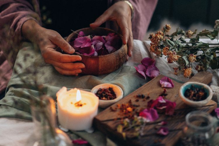 Mãos arrumando pétalas de rosa em tigela de madeira sobre mesa decorada com vela acesa, ervas secas e recipientes com especiarias. Ambiente aconchegante e natural, transmite calor, tranquilidade e conexão com práticas holísticas e espiritualidade. A imagem simboliza a abertura do portal 3/3 para meditação e harmonia espiritual.