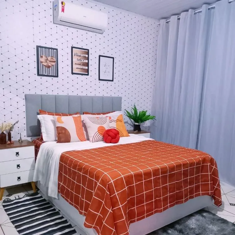 A imagem mostra uma decoração fácil para quarto com uma cama de lençóis brancos e colcha laranja geométrica. Quadros decorativos adornam a parede, um ar-condicionado está acima da cabeceira, e plantas adicionam um toque natural. A combinação de texturas e uso inteligente do espaço criam um ambiente acolhedor e funcional.

