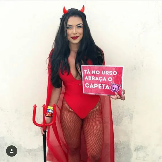 Mulher de pele clara fantasiada de diabinha e placa "Tá no urso abraça o capeta"