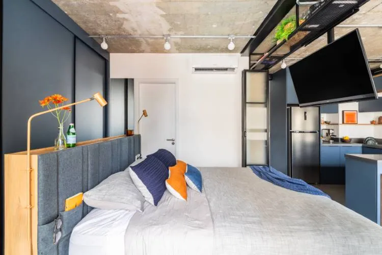 A imagem mostra um quarto moderno com uma cozinha de plano aberto ao fundo. A área do quarto possui uma grande cama com cabeceira estofada cinza, roupa de cama branca e almofadas azul-marinho e laranja. Há uma mesa de cabeceira de madeira com um abajur amarelo visível de um lado da cama. O teto é de concreto, e há uma viga exposta com plantas verdes em cima.

A área da cozinha inclui armários azuis, uma geladeira e o que parece ser um forno ou lava-louças embutido nos armários. As paredes são pintadas de azul escuro ou cinza, contrastando com o piso de madeira clara.