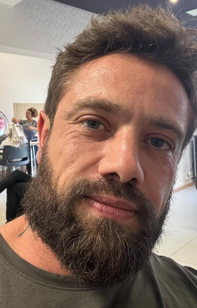 Uma selfie de um homem branco de meia-idade com barba castanha e olhos azuis. Ele sorri para a câmera. Ao fundo, um salão de beleza com uma pessoa sentada na cadeira de cabeleireiro.