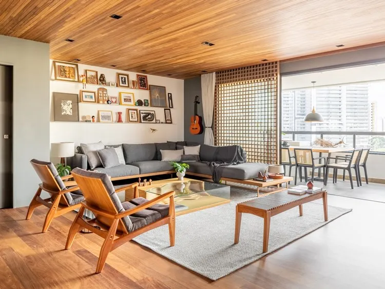 Sala de estar ou living do apartamento decorado de 187 m². Sofá cinza e poltronas com almofadas. Tapete cinza claro, mesa de centro, aparador. Na parede, prateleiras com quadros e outras decoração. Um violão também decorada uma das paredes. A sala de estar é integrada com a varanda, onde tem uma mesa