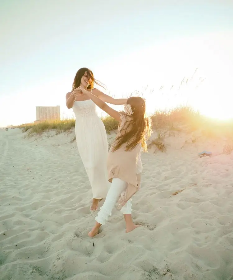 Mãe e filha se divertem na praia ao pôr do sol. Vestindo roupas claras, elas se seguram pelas mãos e giram, rindo. A cena, banhada pela luz dourada, transmite alegria e cumplicidade.