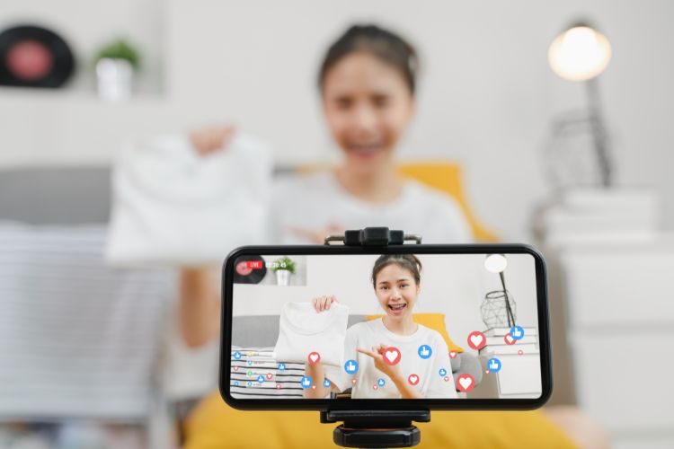 Uma jovem sorridente faz uma live em casa, mostrando uma camiseta branca para a câmera de seu celular em um tripé. A tela do celular mostra a transmissão com ícones de curtidas e corações, sugerindo engajamento da audiência.

