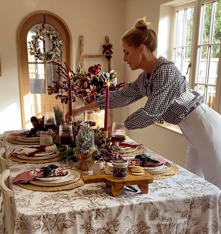Mesa decorada com toalha floral, pratos sobre sousplats de palha, guardanapos vermelhos, taças de vinho, velas vermelhas e arranjo central. Pessoa ajusta a decoração em ambiente iluminado.