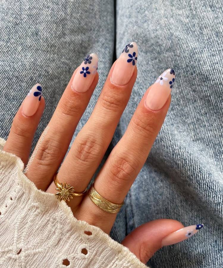 A imagem mostra uma mão com unhas decoradas em esmalte nude, adornadas com pequenas flores azuis nas pontas. Os dedos exibem anéis dourados delicados. O fundo com jeans e uma blusa de tecido bordado complementa o visual casual e elegante.
