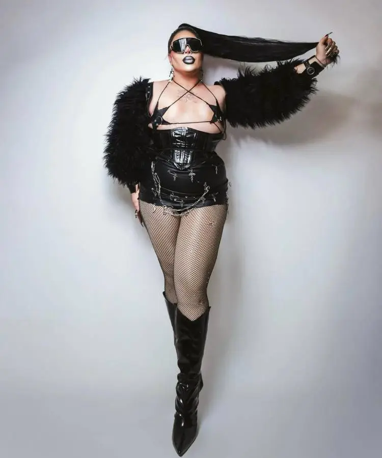 Drag Queen em pé com look preto fetichista: corpete vinil, correntes e meia-arrastão. Óculos escuros grandes e casaco peludo reforçam estética poderosa. Penteado longo preso em rabo de cavalo, botas de cano alto e atitude confiante destacam sensualidade e impacto visual.