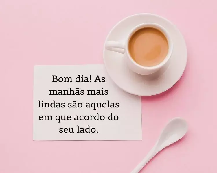 Foto de xícara branca, pires e colher sobre mesa rosa, além de frase: Bom dia! As manhãs mais lindas são aquelas em que acordo do seu lado.