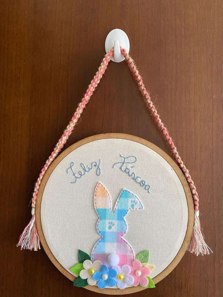 Bastidor de Páscoa bordado "Feliz Páscoa" em azul com um coelho em feltro xadrez colorido e flores. O enfeite está pendurado em uma porta marrom. 