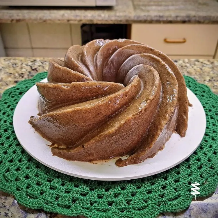 um pudim desenformado em espiral, amarronzado, sobre prato branco sobre sousplat verde de crochê