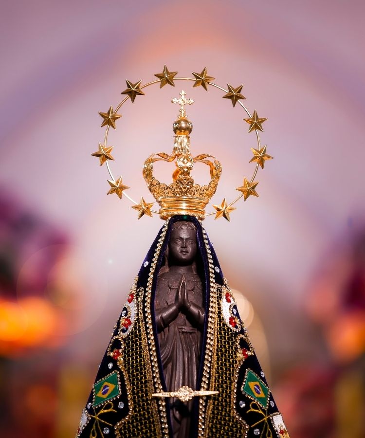 Escultura da Nossa Senhora Aparecida, com manto azul-marinho decorado com as cores da bandeira do Brasil. A santa tem as mãos postas em oração e uma coroa dourada. Estrelas douradas formam um halo ao redor da cabeça.