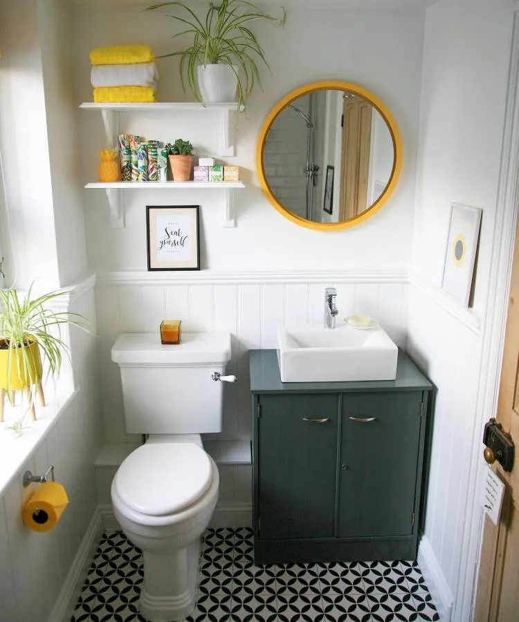 Um banheiro compacto e alegre com piso de azulejos preto e branco. Possui um vaso sanitário branco, um gabinete cinza-escuro com pia de bancada branca, e um espelho redondo com moldura amarela. Prateleiras brancas com plantas e toalhas amarelas decoram o ambiente.

