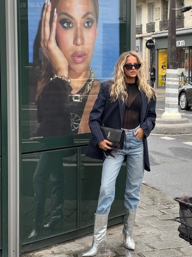 mulher usando body de renda preto com calça jeans azul, blazer azul risca de giz, bota de salto metalizada e bolsa preta