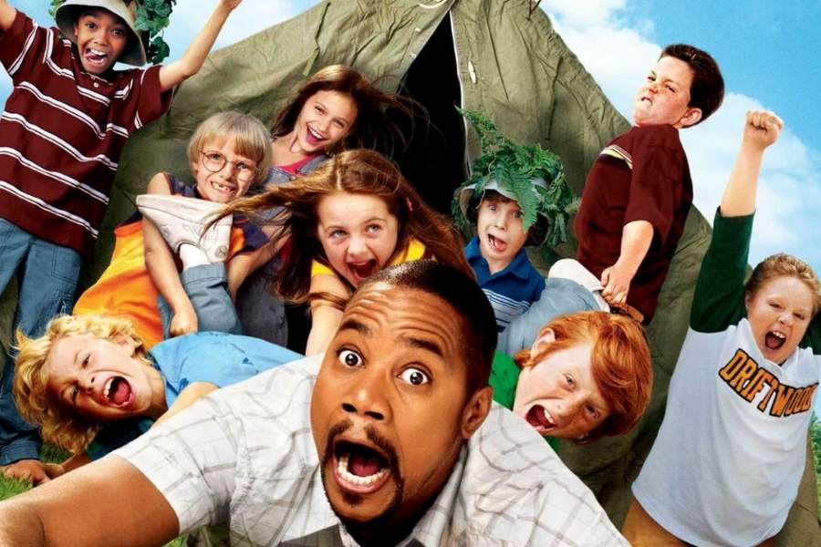 Foto do personagem Charlie Hinton (Cuba Gooding Jr.)deitado no chão com várias crianças ao seu redor. No fundo, uma barraca montada e o céu azul com nuvens.