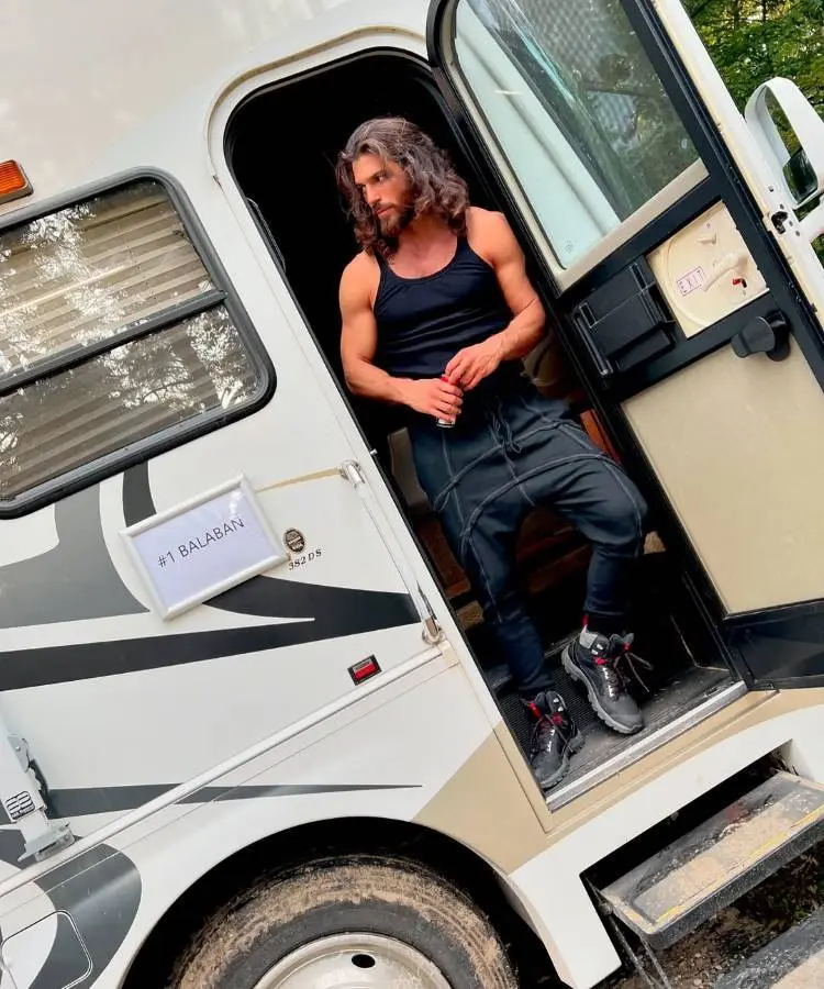 Homem de cabelos longos e barba, vestindo regata preta e calça esportiva, está na porta de um trailer branco. Ele olha para o lado com expressão pensativa. A placa na porta indica "#1 Balaban".