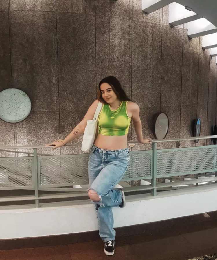 Imagem com fundo de grade. No centro, mulher usando cropped regata verde e amarelo, calça jeans, tênis vans e bolsa 
