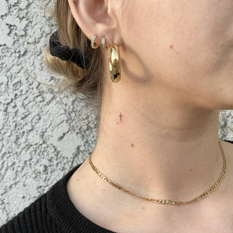 A imagem mostra uma pessoa de perfil com cabelo preso e brincos dourados. Ela tem uma tatuagem no pescoço de uma pequena cruz preta, localizada logo abaixo da orelha. A tatuagem é discreta e minimalista. A pessoa também usa uma corrente dourada fina e uma camiseta preta.




