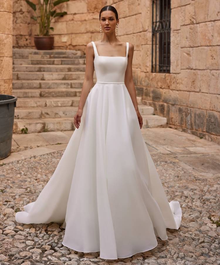 Noiva em vestido branco clássico com decote quadrado, alças finas e saia volumosa fluida. O design minimalista destaca elegância e sofisticação. Ao fundo, escadaria de pedra e parede rústica criam um cenário atemporal e encantador.





