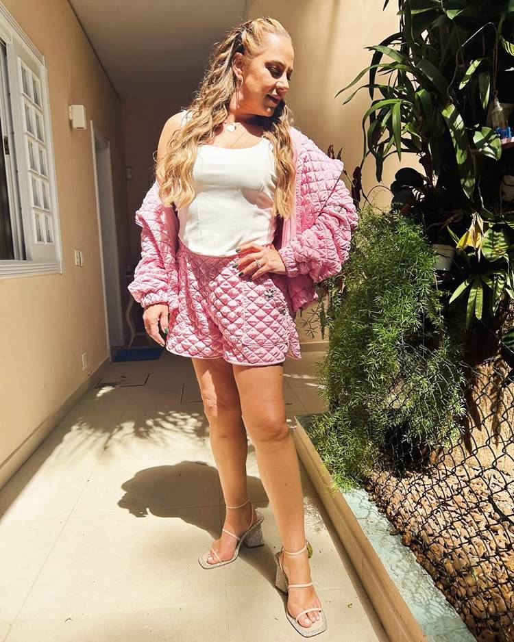 Silvia Abravanel com look branco e rosa em um corredor de casa com janelas porta e plantas