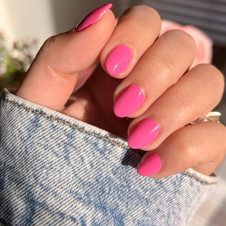 Mão feminina com unhas curtas e ovais pintadas em rosa vibrante, com acabamento brilhante. A manga de jeans desbotado adiciona textura casual ao fundo claro, destacando o tom alegre e feminino, na tendência de esmaltes rosa candy para 2025.