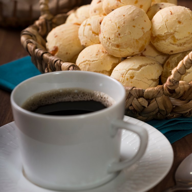 Xícara com café e pires e cesta de pão de queijo ao fundo