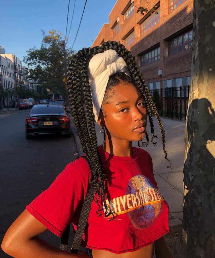 Mulher com tranças longas, lenço branco enrolado no topo da cabeça, camiseta vermelha e bolsa preta, posando em rua urbana ao pôr do sol.