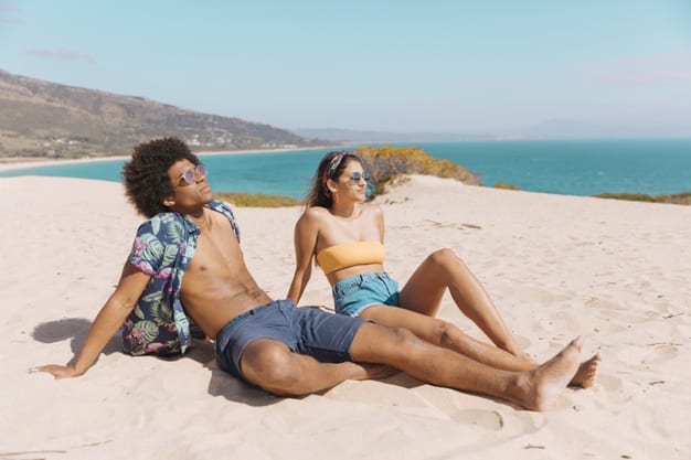 Homem de pele negra e mulher de pele clara tomando sol na areia de uma praia. 