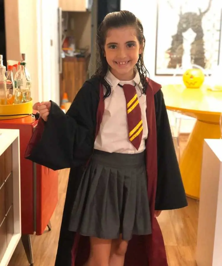 Garota com fantasia de bruxa infantil Harry Potter. Cores predominantes: branco, preto, vinho, amarelo, cinza. Usa: Camiseta, saia, capa longa com touca, gravata