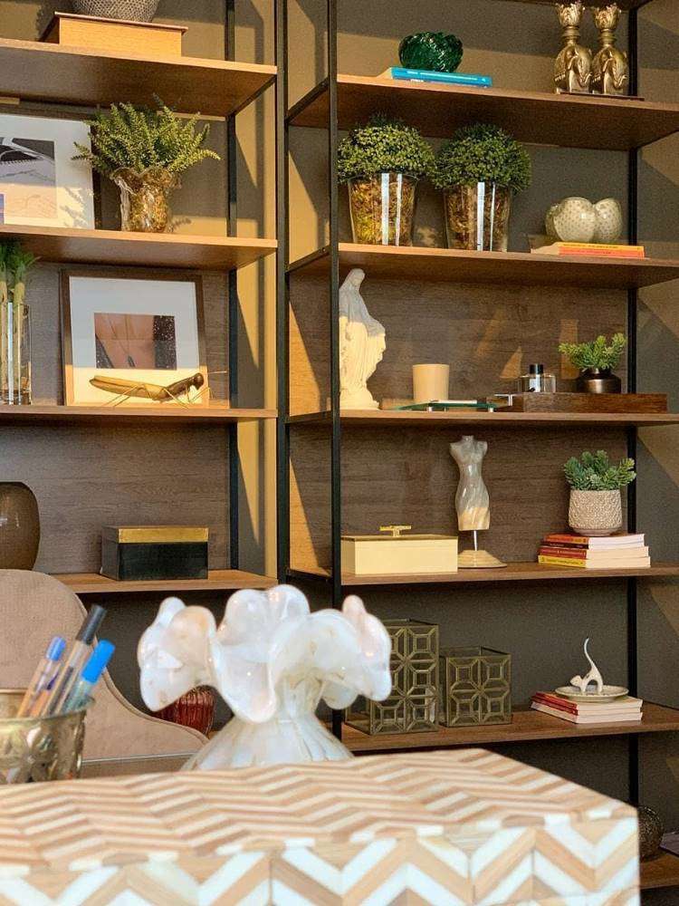 Estante de madeira e metal, com prateleiras abertas. Nela, há vasos de plantas, quadros, livros, diversos itens decorativos e uma vela circular. 