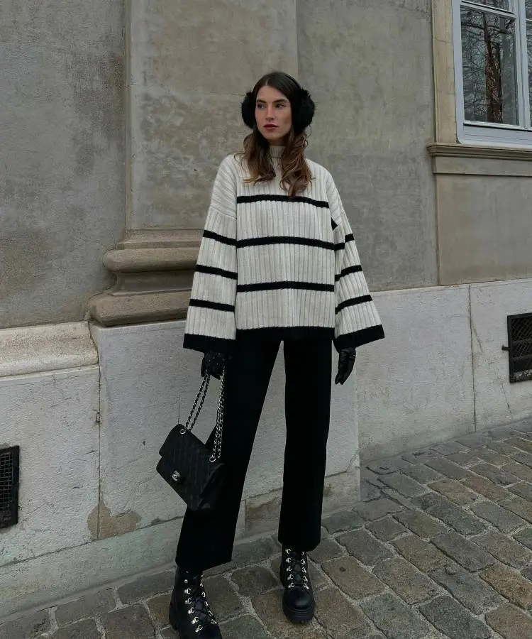 Uma mulher com cabelo castanho liso usa um suéter de tricô listrado em creme e preto, calças pretas e botas pretas de cadarço. Ela completa o look com protetores de orelha pretos e luvas pretas, segurando uma bolsa preta acolchoada com corrente.