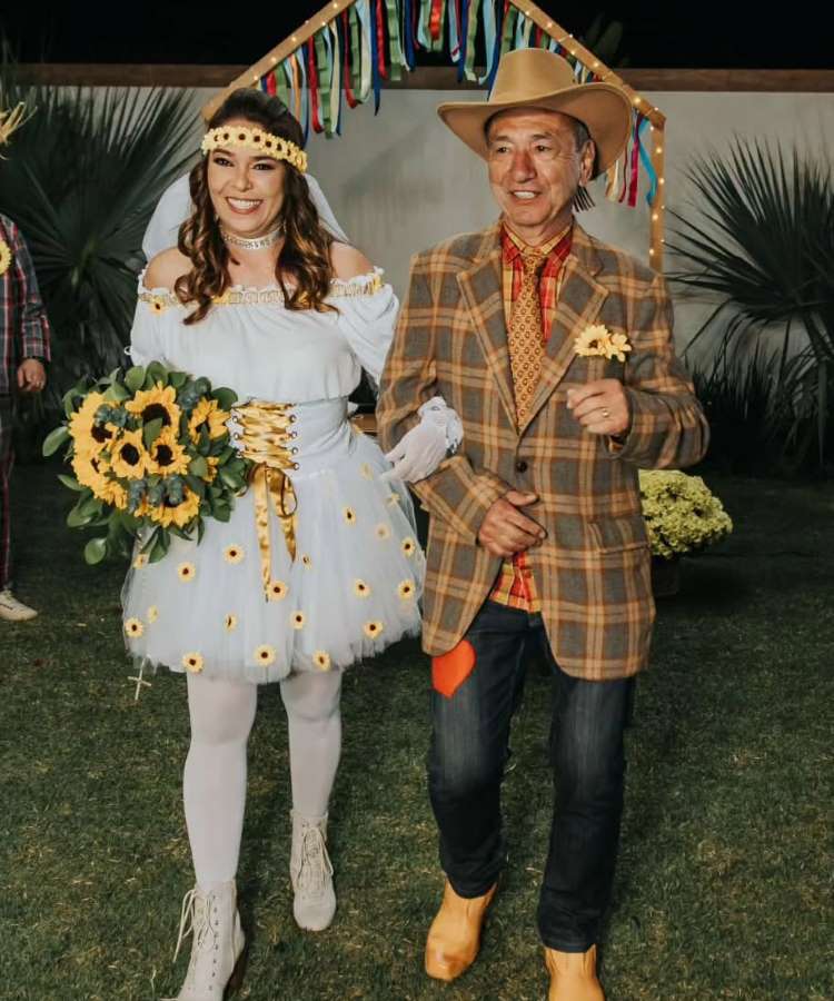 Uma noiva em um vestido branco decorado com girassóis e véu caminha de braço dado com um homem de terno xadrez e chapéu de cowboy. Ela segura um buquê de girassóis. O cenário ao ar livre com gramado e um arco decorativo com fitas coloridas sugere um casamento festivo ou temático de Festa Junina.