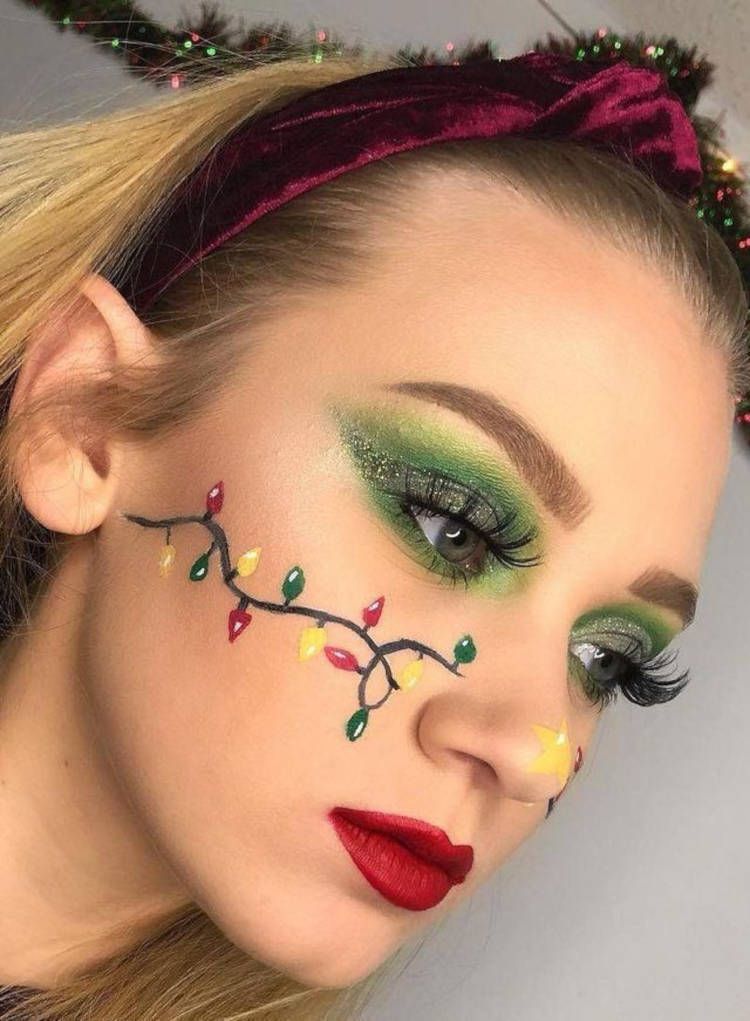 Mulher de pele clara com sombra verde, batom vermelho, estrela amarela na ponta do nariz e desenho de luzes de Natal na bochecha 