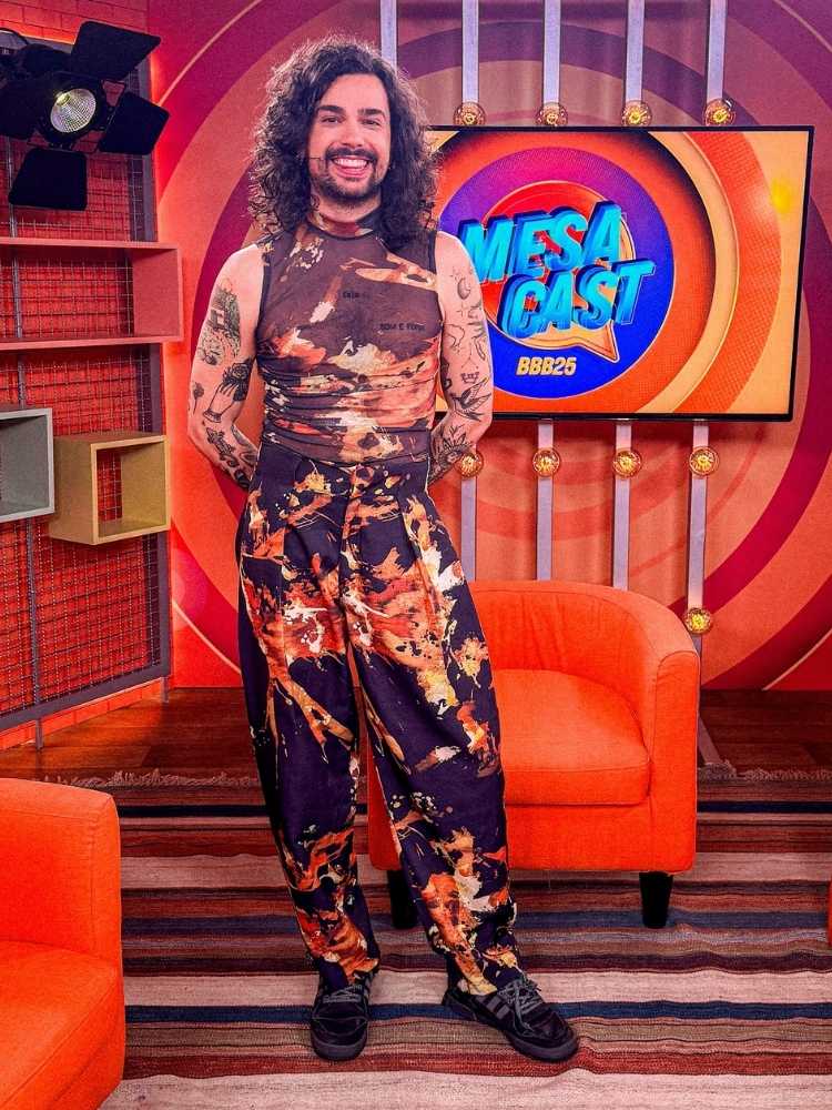 Vitor DiCastro com macacão marrom e laranja no cenário do MesaCast BBB