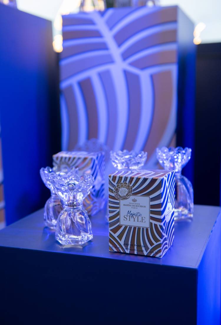 Frascos de perfume com tampas em forma de flor estão sobre uma superfície azul. Ao lado, uma caixa com o rótulo "Majestic Style". O fundo exibe padrões geométricos em azul e branco, criando uma atmosfera elegante e moderna.





