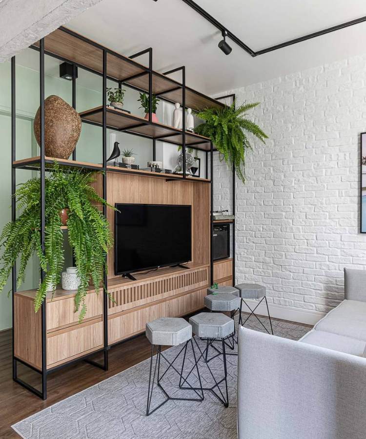 Estante de madeira e ferragem com decorações, TV e dois vasos de planta samambaia. Ao lado, bancos, sofá e tapete