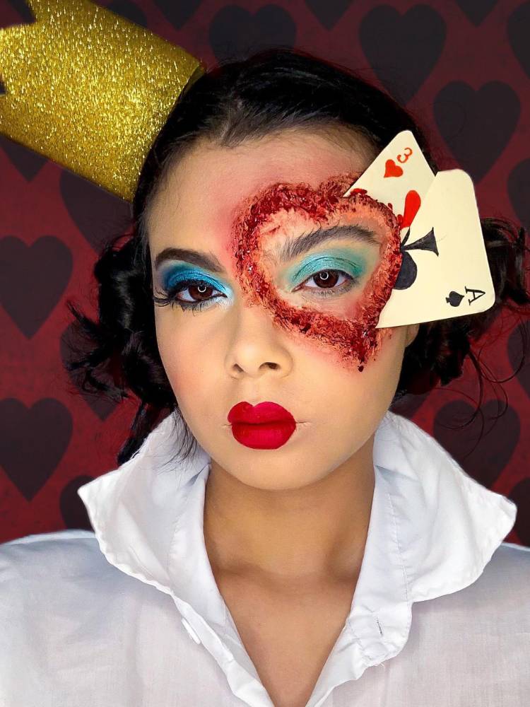 Mulher de pele clara fantasiada de Rainha de Copas com machucado em formato de coração com cartas no ferimento com sangue fake