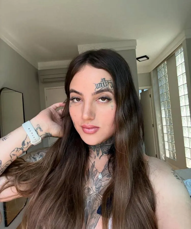 Retrato de jovem de cabelos longos castanhos, exibindo tatuagens no pescoço, braço e testa. Ela veste blusa branca cavada e usa relógio no pulso. Pose confiante com a mão no cabelo, em ambiente moderno com paredes claras e vidro.