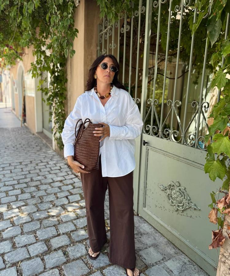 Uma mulher de óculos escuros, blusa social branca e calças marrom largas. Ela segura uma bolsa de palha trançada, em pé na frente de um portão de ferro, em uma rua de paralelepípedos com vegetação.