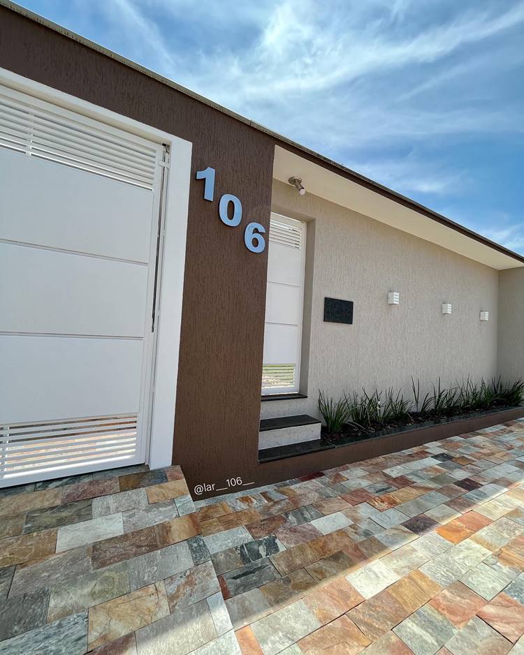 Muro com textura marrom e cinza-claro, com portão branco, número 106 e luminárias