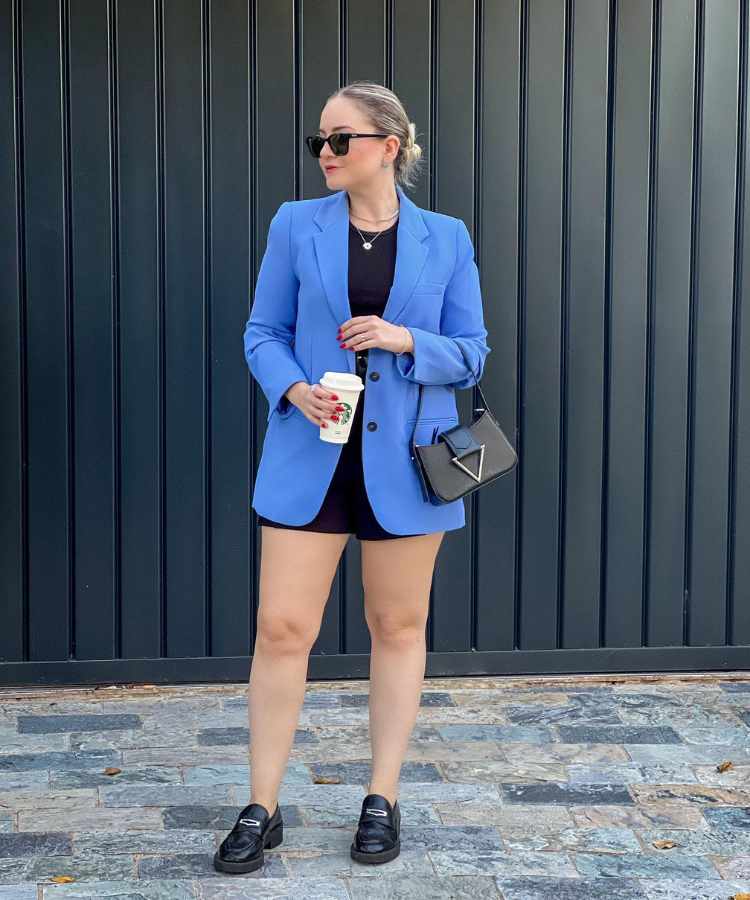 Imagem com fundo de portão preto. No centro, mulher usando camisa preta, short preto, blazer azul, bolsa preta, óculos preto e mocassim preto