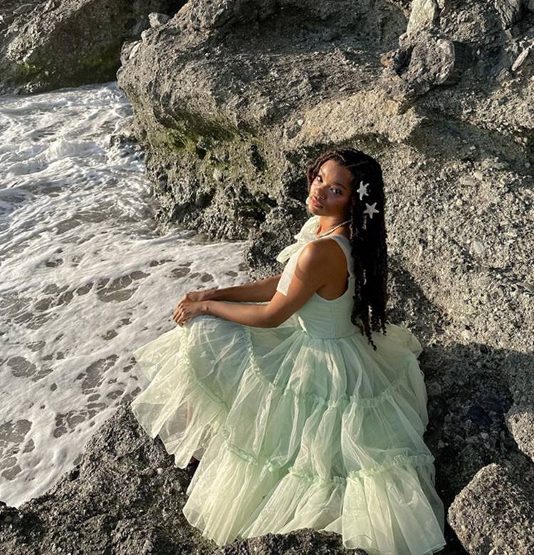Menina com vestido babado e penteado com acessórios de estrela sentada em pedras no mar

