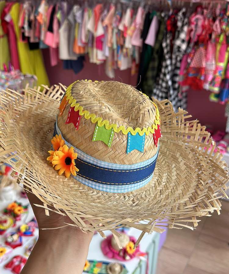 Uma mão segura um chapéu de palha decorado com bandeirinhas coloridas, uma fita azul e laranja e pequenas flores alaranjadas. Ao fundo, peças de vestuário coloridas, possivelmente roupas de festa junina, estão penduradas em araras.