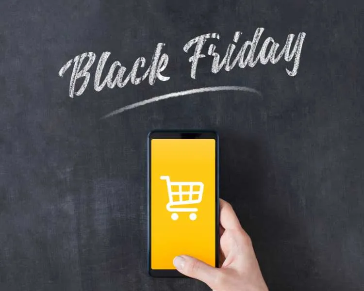 Mão segurando celular que tem desenho de carrinho de compras em fundo amarelo. No fundo da foto, quadro negro e escrito Black Friday