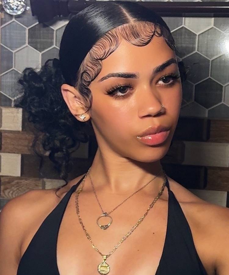 Mulher com cabelo preso em coque baixo, baby hairs estilizados em curvas elegantes na testa. Ela usa maquiagem leve, brincos pequenos, colares dourados delicados e top preto, posando em ambiente com fundo de azulejos hexagonais em tons neutros.