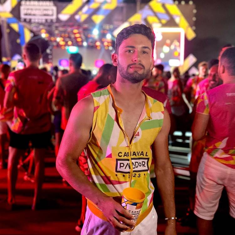 A imagem mostra um homem em uma festa noturna de Carnaval, usando um abadá customizado colorido com estampas geométricas em amarelo, verde e rosa. Ele segura um copo estilizado, cercado por pessoas em um ambiente iluminado vibrante.