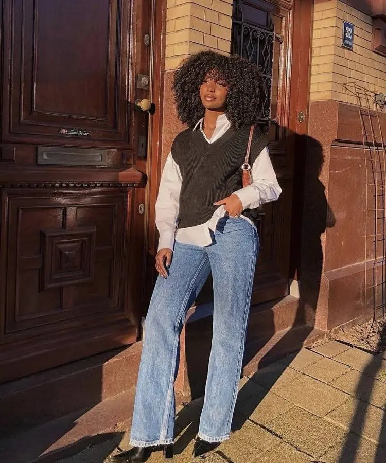 Uma mulher com cabelo encaracolado e volumoso veste um colete de tricô preto sobre uma camisa branca de botões, combinando com jeans largos de lavagem clara e sapatos pretos de bico fino. Ela posa em frente a uma porta de madeira escura, com luz solar criando sombras.