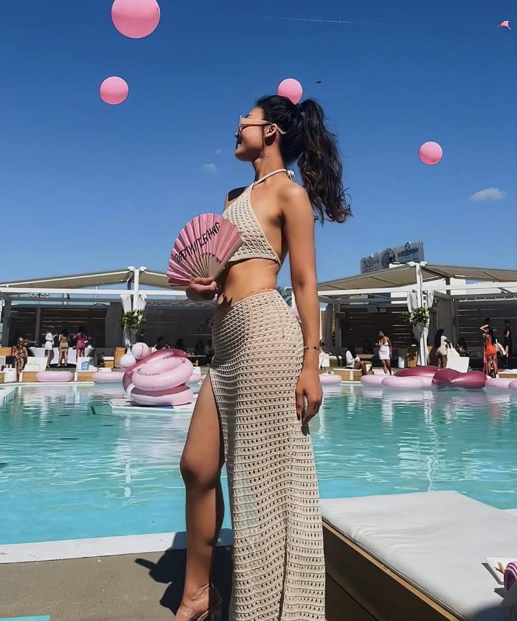 Mulher com rabo de cavalo e óculos de sol, segurando um leque rosa. Ela usa uma saída de praia de crochê longa e bege com uma fenda. Ao fundo, uma piscina com boias e balões rosa.